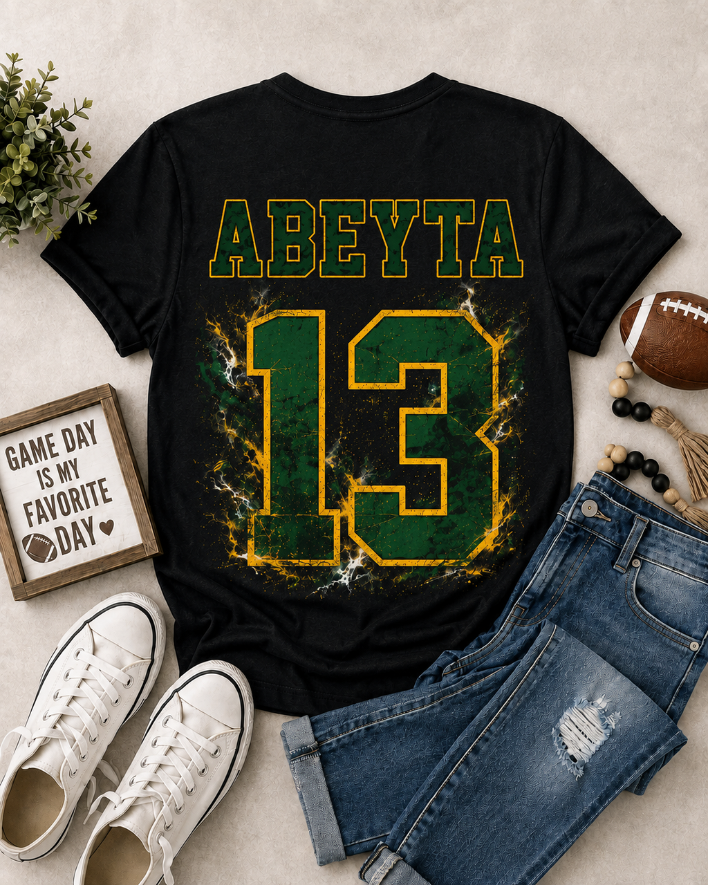 A black short-sleeve graphic T-shirt laid flat on a light beige surface, styled with blue jeans, white sneakers, a football, beads, and a small sign. The shirt has the name 'ABEYTA' in green outlined yellow letters above a large distressed green number 13 with yellow edging and white crackle effects.