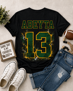 A black short-sleeve graphic T-shirt laid flat on a light beige surface, styled with blue jeans, white sneakers, a football, beads, and a small sign. The shirt has the name 'ABEYTA' in green outlined yellow letters above a large distressed green number 13 with yellow edging and white crackle effects.
