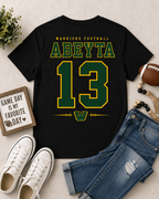A black graphic t-shirt featuring 'ABEYTIA 13' in green and yellow varsity lettering, styled in a flat lay with blue jeans, white sneakers, a football, and a decorative sign on a light beige background.