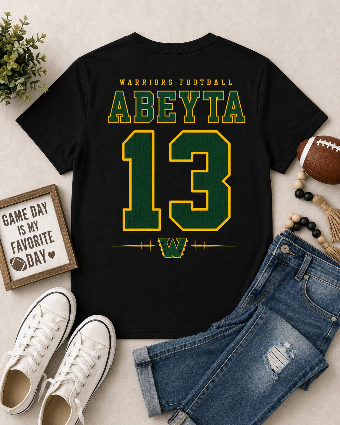 A black graphic t-shirt featuring 'ABEYTIA 13' in green and yellow varsity lettering, styled in a flat lay with blue jeans, white sneakers, a football, and a decorative sign on a light beige background.