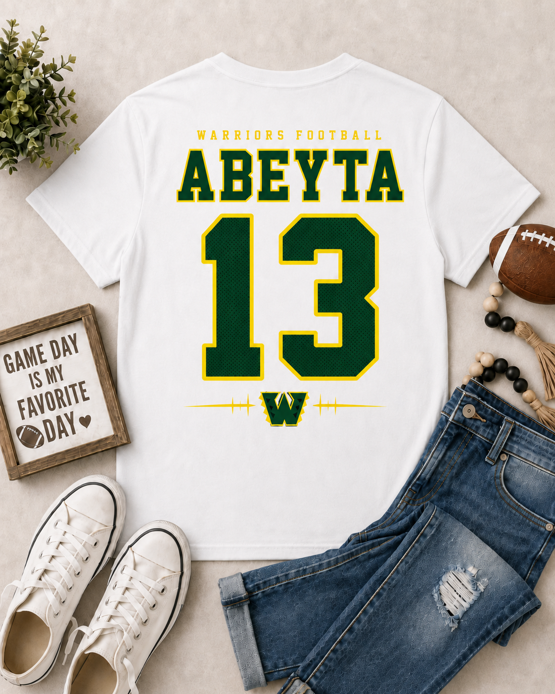 A white graphic t-shirt featuring 'ABEYT A 13' and 'WARRIORS FOOTBALL' in green and yellow, styled in a flat lay with blue jeans, white sneakers, a football, and a small framed sign on a beige background.