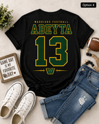 A black short-sleeve graphic T-shirt laid flat with jeans, white sneakers, a football, beads, and a small sign around it on a light neutral surface. The shirt reads 'WARRIORS FOOTBALL', 'ABEYTA', and '13' in green and yellow, with a 'W' logo at the bottom.