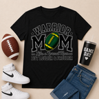 A black graphic t-shirt featuring a football-themed design with the words 'WARRIOR MOM' and 'Like a Normal Mom BUT LOUDER & PROUDER'. The t-shirt is styled in a flat-lay arrangement with a brown football, white Nike high-top sneakers, blue jeans, and a black insulated tumbler with a straw on a light beige background.
