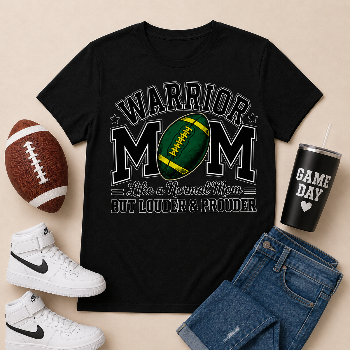 A black graphic t-shirt featuring a football-themed design with the words 'WARRIOR MOM' and 'Like a Normal Mom BUT LOUDER & PROUDER'. The t-shirt is styled in a flat-lay arrangement with a brown football, white Nike high-top sneakers, blue jeans, and a black insulated tumbler with a straw on a light beige background.