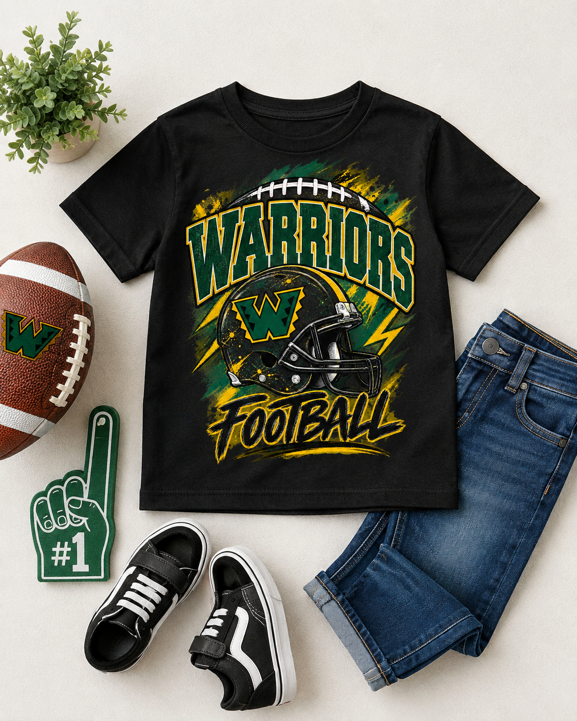 A black kids' short-sleeve T-shirt with a large 'WARRIORS FOOTBALL' graphic in green, yellow, and white, styled in a flat lay with a football, blue jeans, black sneakers, a green foam finger, and a small potted plant on a light beige background.