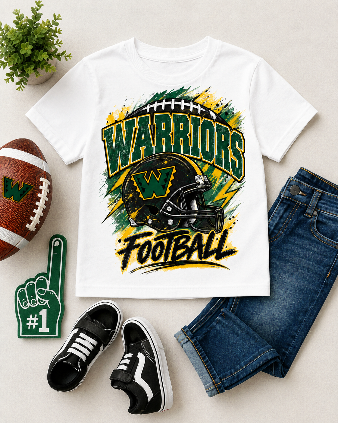 A white kids' short-sleeve T-shirt with a green, yellow, and black 'Warriors Football' graphic, styled in a flat lay with blue jeans, black sneakers, a football, a foam finger, and a small plant on a light beige background.