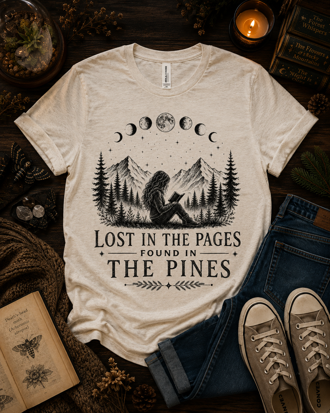 A cream-colored graphic t-shirt laid flat on a dark wood background, surrounded by boho-style props including blue jeans, canvas sneakers, an open book, a candle, and a small terrarium. The t-shirt features a black graphic of a person reading in a pine forest with moon phases above, and the text 'LOST IN THE PAGES FOUND IN THE PINES'.