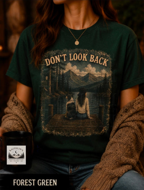 A woman wearing a forest green graphic t-shirt with a mountain lake scene and the text 'DON'T LOOK BACK'. She has a brown knit cardigan draped off her shoulders, and a candle in the foreground labeled 'CANDLE CO' with 'FOREST GREEN' text at the bottom.