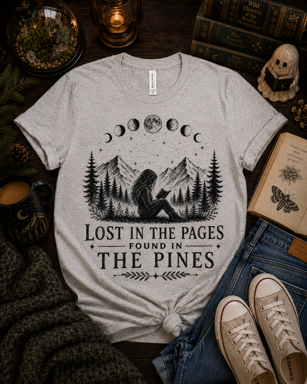 A light heather gray graphic t-shirt featuring a black illustration of a girl reading in a pine forest under a moon phase sky, with the text 'LOST IN THE PAGES FOUND IN THE PINES'. The t-shirt is styled in a cozy flat-lay arrangement with blue jeans, beige sneakers, a knit sweater, an open book, and a mug on a dark wooden background.