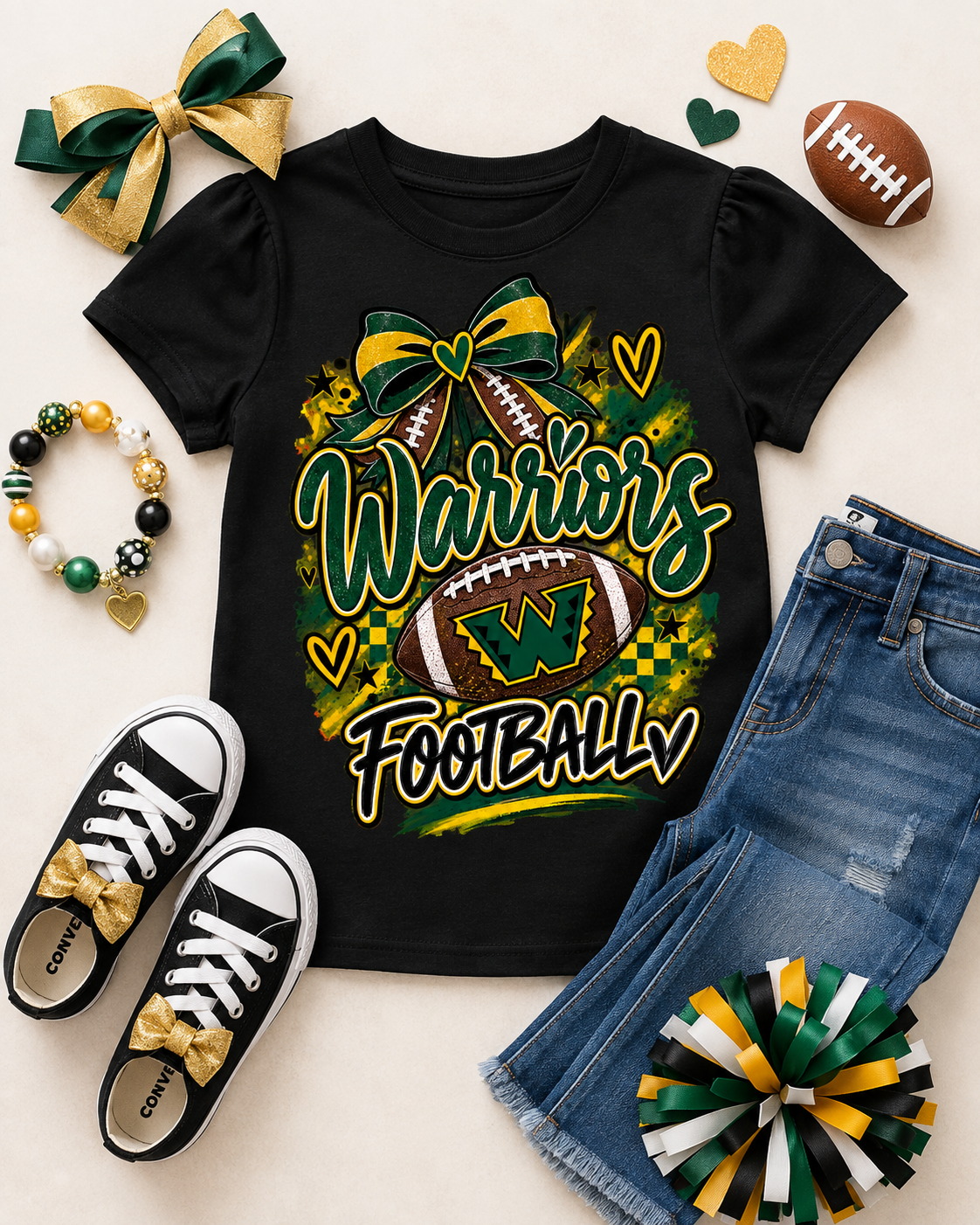 A black short-sleeve graphic T-shirt laid flat with jeans, black sneakers, a green-and-gold bow, a football, heart-shaped accessories, and a pom-pom arranged around it. The shirt graphic reads 'Warriors Football' with a football and bow design in green, yellow, white, and black.