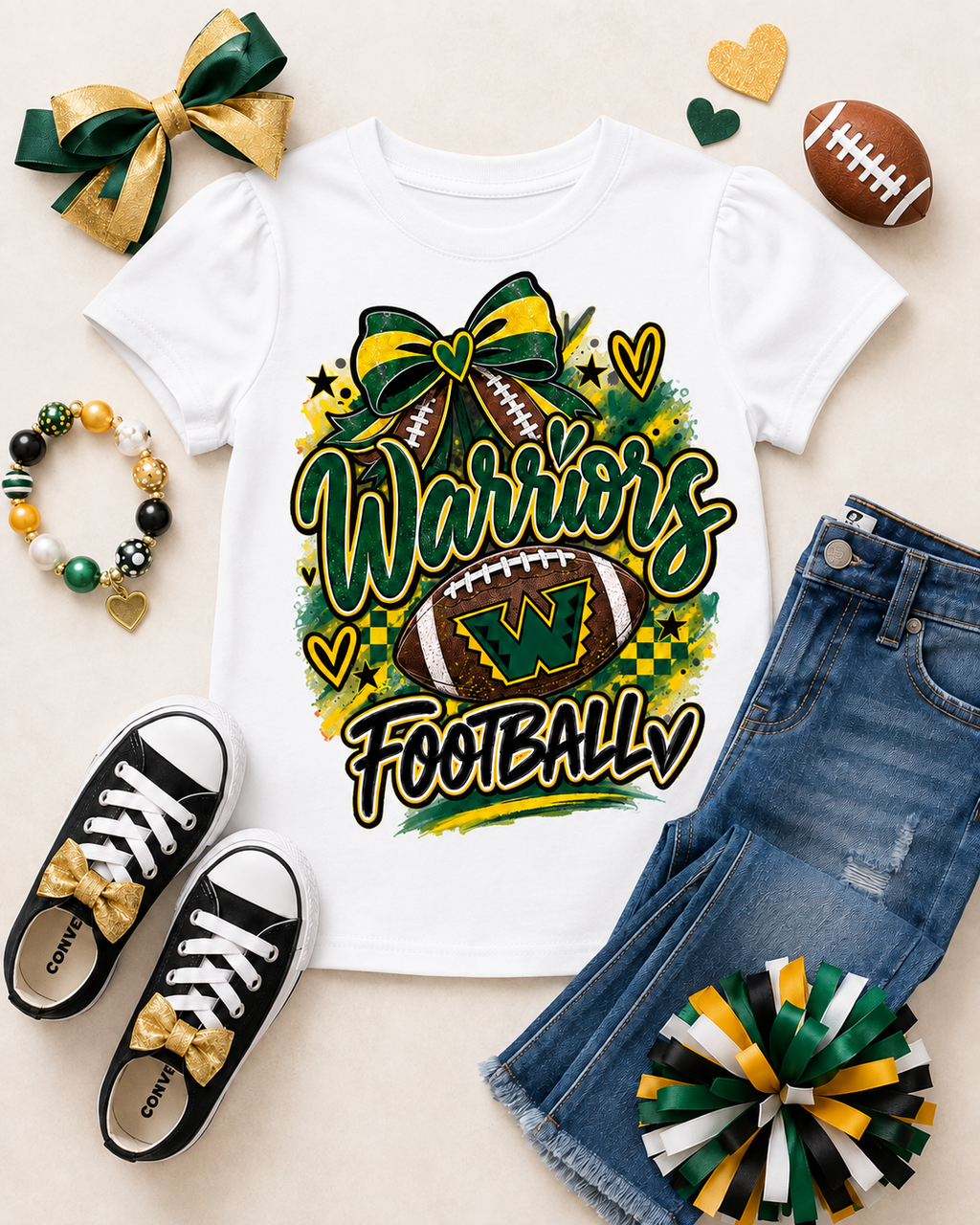 A white short-sleeve graphic T-shirt laid flat with a colorful 'Warriors Football' design featuring a football, bow, hearts, and checkered accents. The shirt is styled in a flat lay with blue jeans, black sneakers with gold bows, a green and gold ribbon, a beaded bracelet, a football, and heart-shaped decorations on a light beige background.
