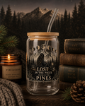 A clear glass tumbler with a bamboo lid and glass straw, printed with a dark forest and mountain illustration and the phrase 'LOST IN THE PAGES FOUND IN THE PINES.' It is styled on a rustic wooden surface with books, a candle, pine branches, and a knit blanket against a moody mountain-and-tree background.