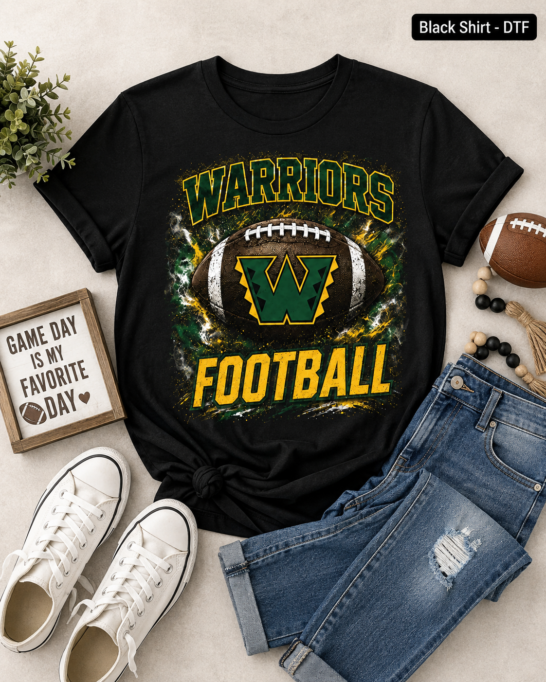 A black graphic t-shirt featuring a football design with the text 'WARRIORS FOOTBALL' and a large green 'W' logo, styled in a flat lay with blue jeans, white sneakers, a small sign, and a football on a light beige background.