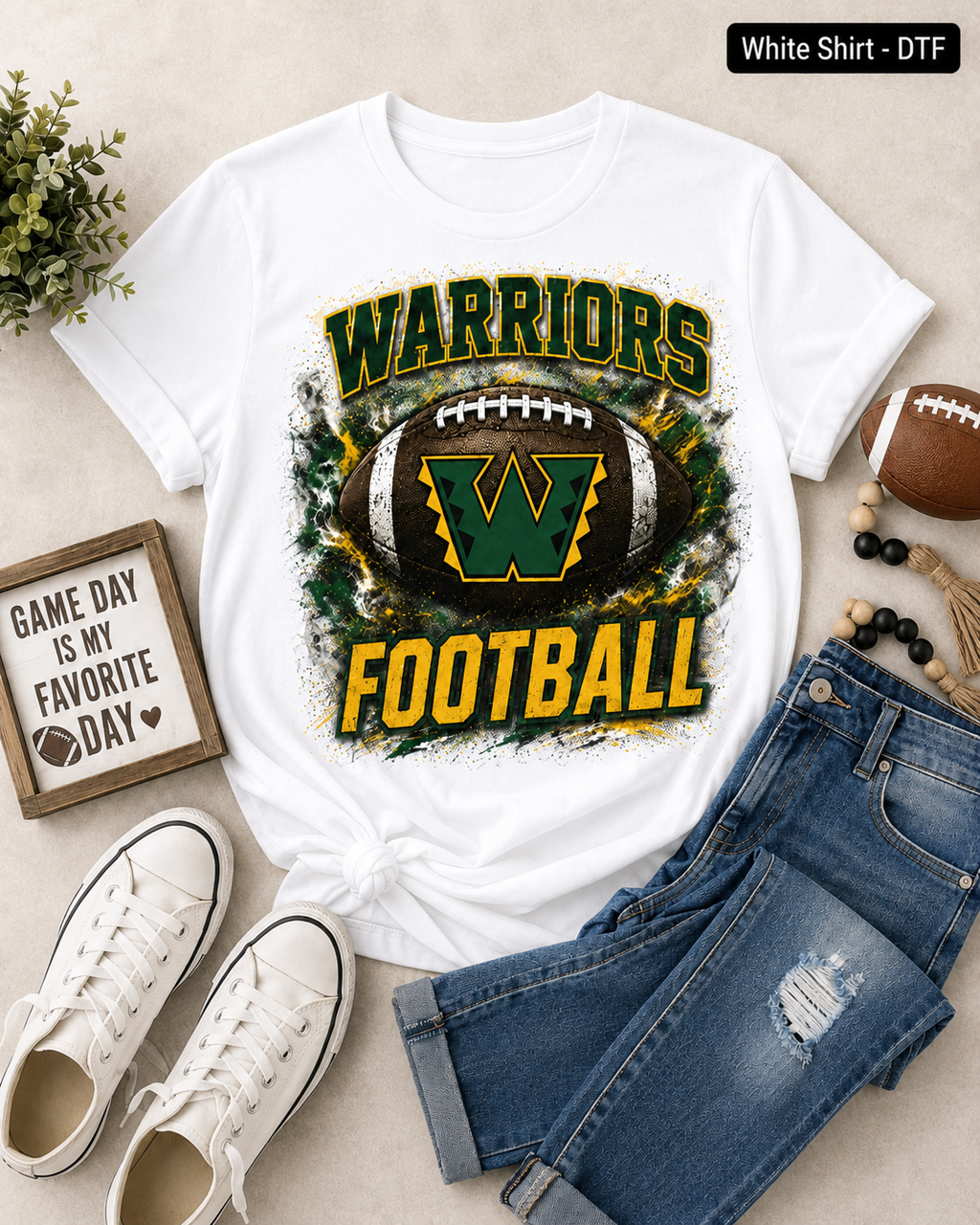 A white graphic t-shirt featuring a distressed green and yellow 'WARRIORS FOOTBALL' design with a football and 'W' logo, styled in a flat lay with blue jeans, white sneakers, a small sign, and a football on a beige background.