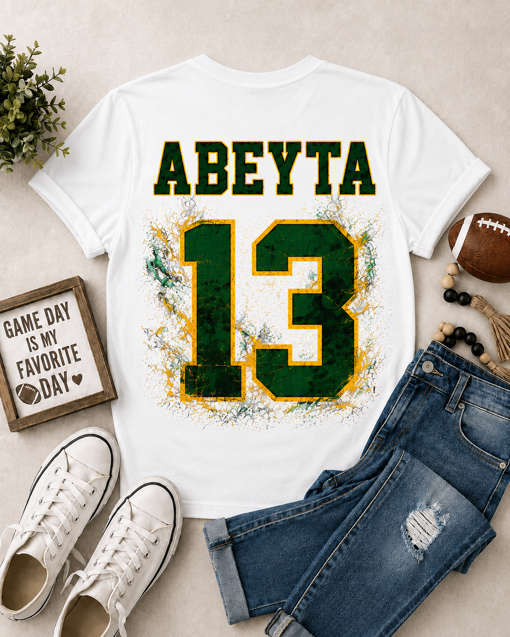 A white graphic T-shirt laid flat on a light beige surface, styled with blue jeans, white sneakers, a football, beads, and a small sign. The shirt has the name 'ABEYTta' in green outlined letters above a large distressed green number 13 with yellow/gold and white splatter details.