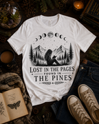 A white graphic t-shirt with a black moon phase and mountain illustration, styled in a cozy flat-lay with jeans, sneakers, a book, and a candle on a dark wooden surface.
