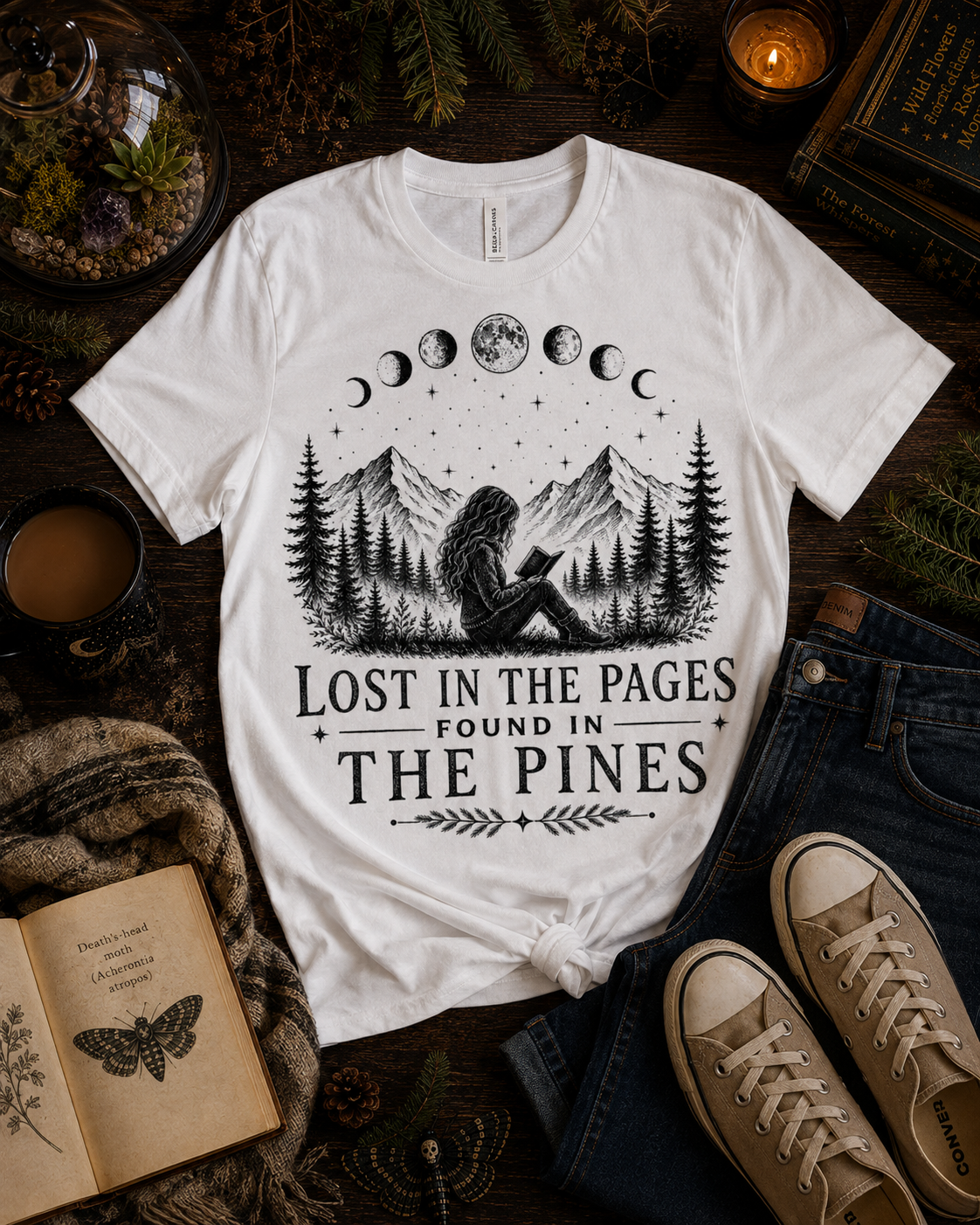 A white graphic t-shirt with a black moon phase and mountain illustration, styled in a cozy flat-lay with jeans, sneakers, a book, and a candle on a dark wooden surface.