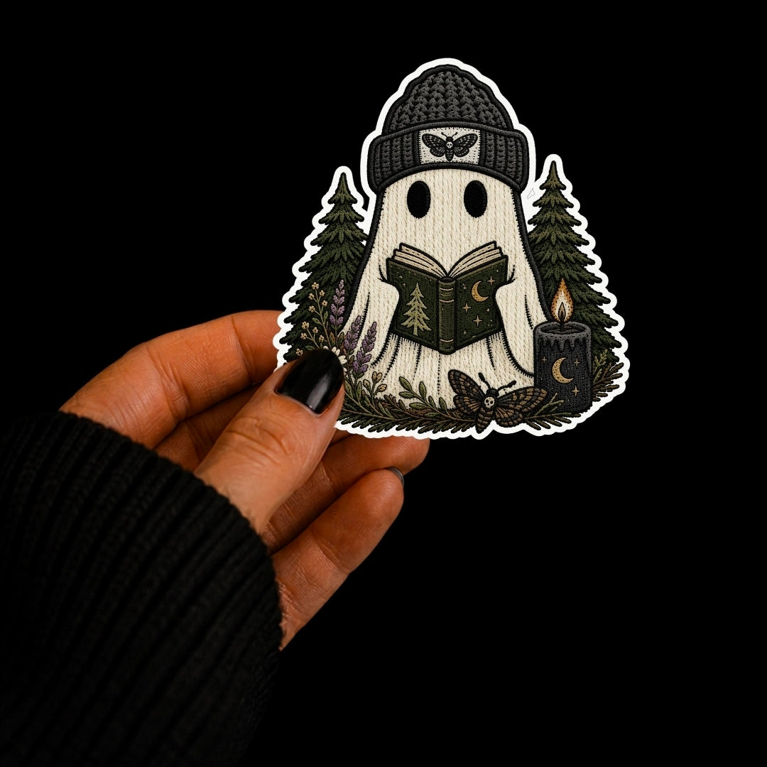 A hand with black nail polish holds a die-cut sticker featuring a ghost reading a book in a forest, set against a solid black background.