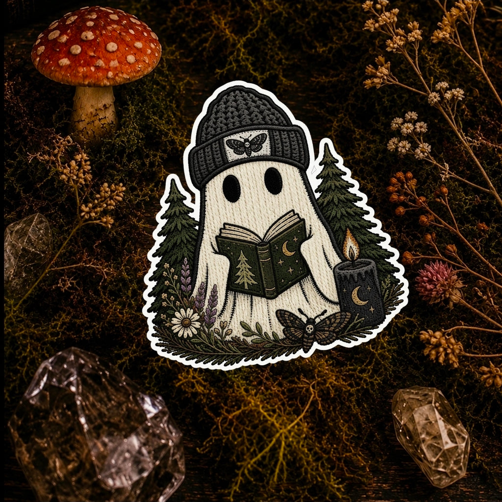 A ghost-shaped embroidered patch or sticker featuring a white ghost wearing a black knit beanie and reading a book, surrounded by trees, flowers, a candle, and a butterfly, displayed on a dark mossy background with a red mushroom and crystals.