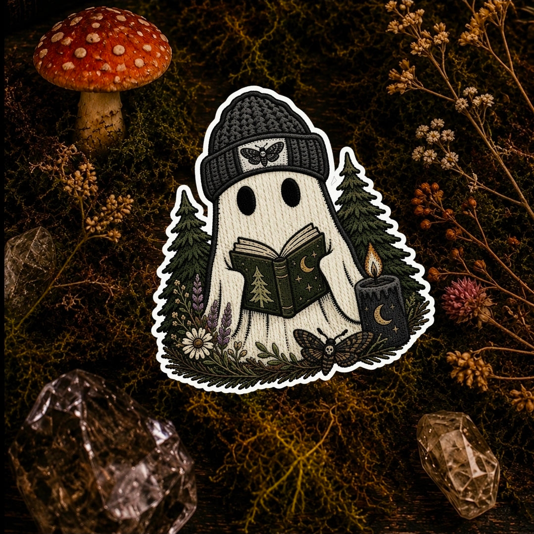 A ghost-shaped embroidered patch or sticker featuring a white ghost wearing a black knit beanie and reading a book, surrounded by trees, flowers, a candle, and a butterfly, displayed on a dark mossy background with a red mushroom and crystals.