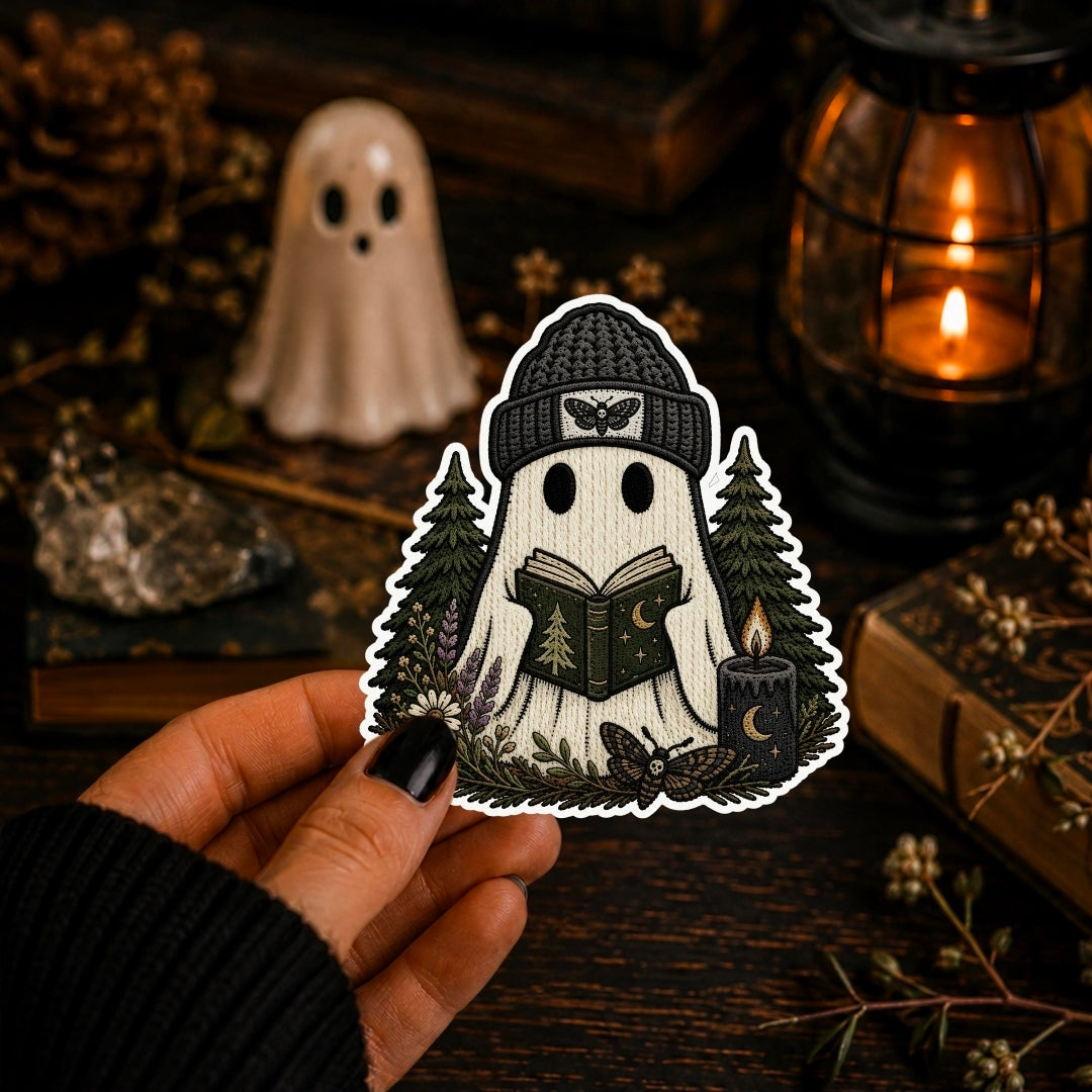 A hand holds a die-cut sticker featuring a cute ghost character reading a book in a forest, surrounded by trees, a candle, and a butterfly. The background is a cozy, dark, autumnal scene with a lit lantern, books, and a small ghost figurine.