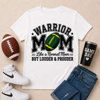 A white graphic T-shirt laid flat on a beige background, surrounded by a brown football, white high-top sneakers, blue jeans, and a black tumbler with the words 'GAME DAY'. The shirt reads 'WARRIOR MOM Like a Normal Mom BUT LOUDER & PROUDER' with a green and yellow football graphic.